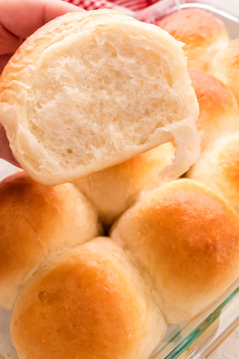 Close-up of a homemade dinner roll showing the fluffy inside over a pan of rolls just out of the oven.
