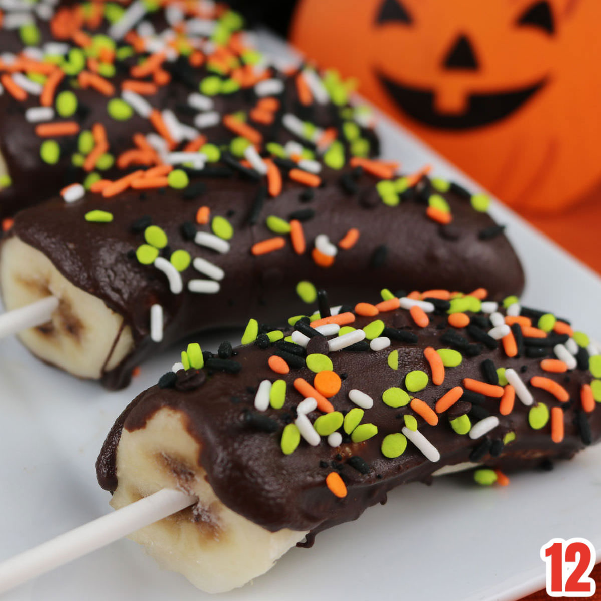 Close up of four Halloween Frozen Bananas laying on a white platter in front of a Orange Jack-o-Lantern Halloween decoration.