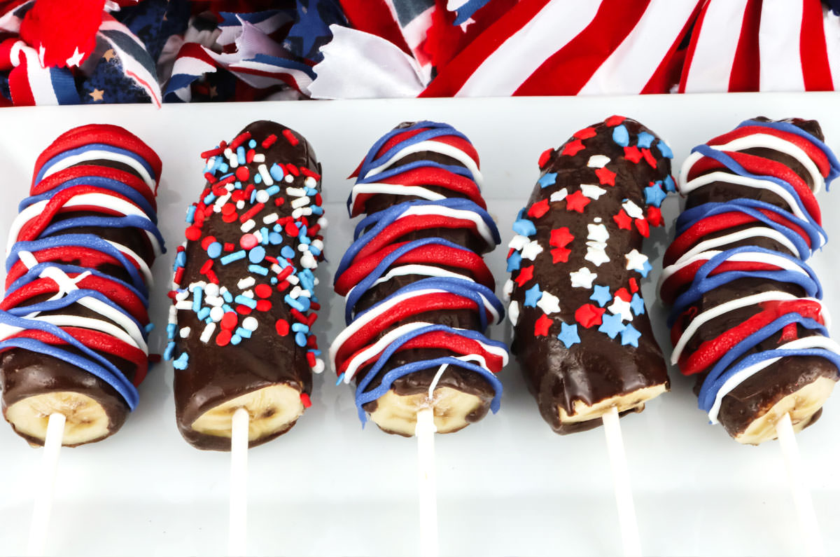 Closeup on five Frozen Chocolate-Covered Bananas laying on a white platter and decorated in Red White and Blue for 4th of July.
