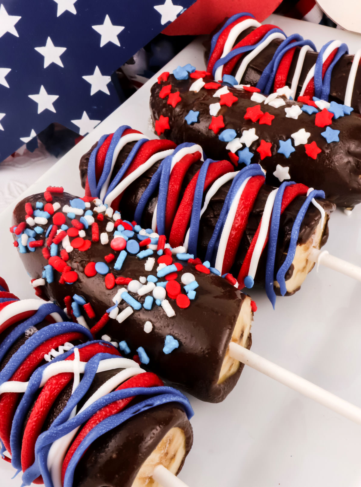 Close up on five Frozen Chocolate-Covered Bananas laying on a white serving platter in from of Red White and Blue 4th of July decorations.
