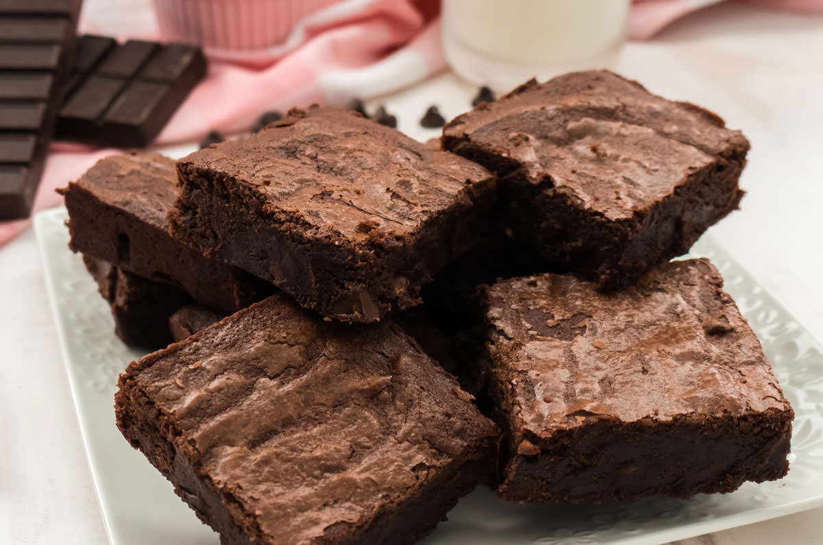 Close up of a plate of Homemade Brownies in front of a chocolate bar, a glass of milk and a pink table linen.