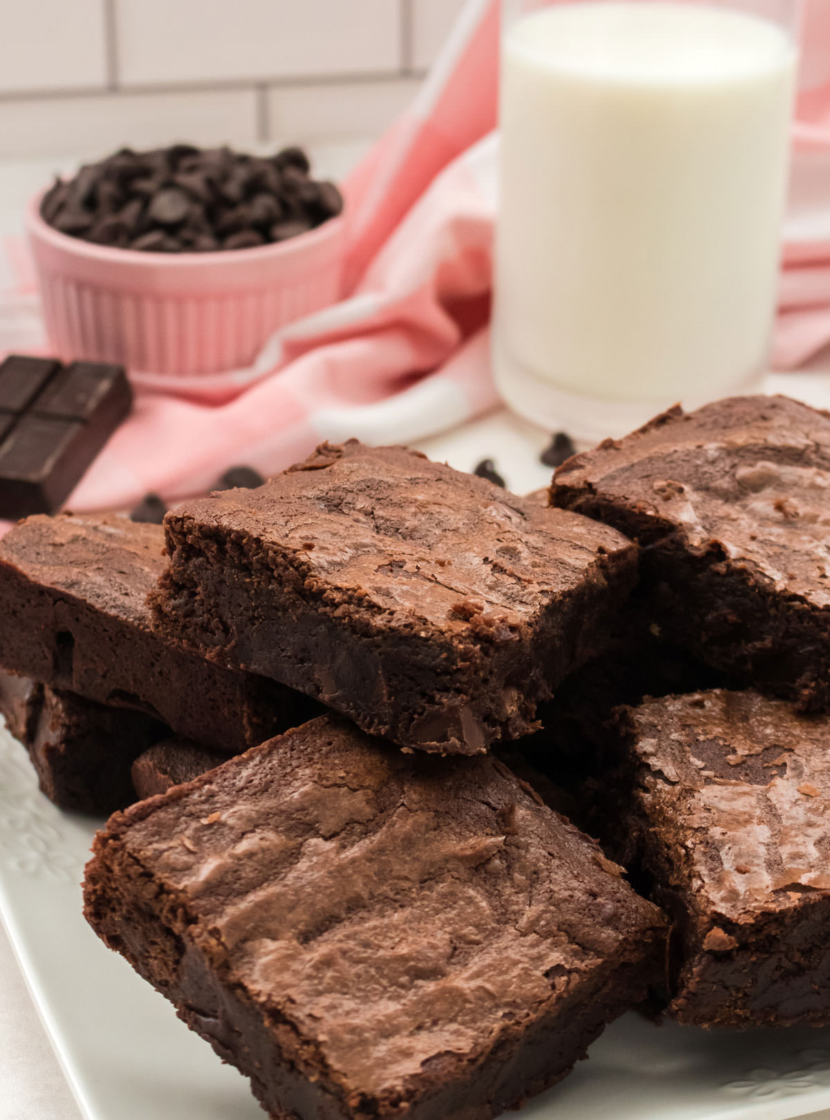 Close up on a white plate filled with Homemade Fudge Brownies along with a glass of milk and a ramekin of chocolate chips in the background.