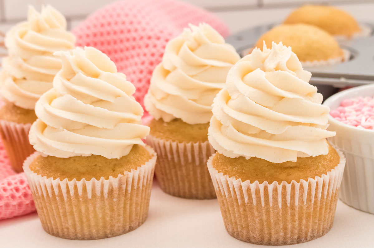 Closeup on four Homemade Vanilla Cupcakes sitting on a white table in front of a pink table linen, a ramekin filled with pink sprinkles and a cupcake pan.