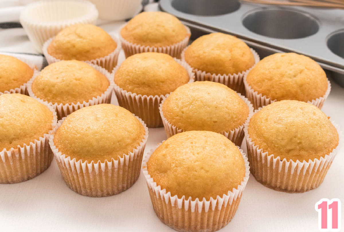 Closeup on a dozen yellow cupcakes in white cupcake liners sitting in front of a cupcake tin and white cupcake liners.