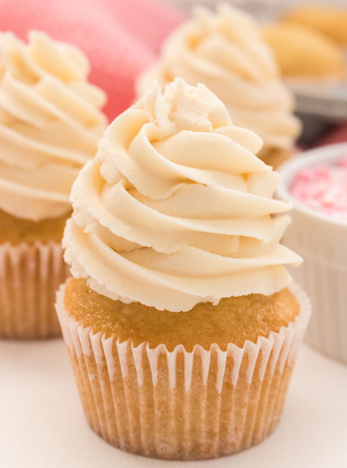 Closeup on a Homemade Vanilla Cupcake topped with Buttercream Frosting sitting next to a ramekin full of pink sprinkles and more yellow cupcakes.