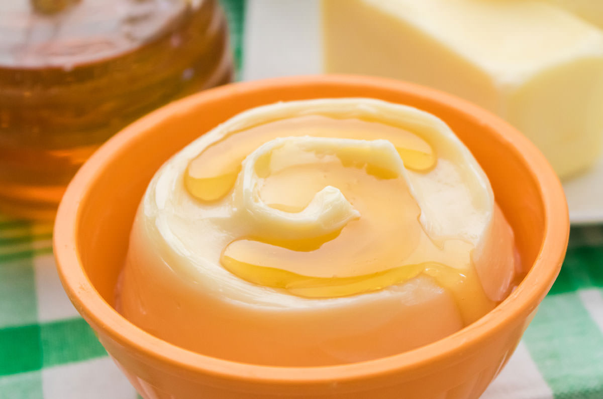 Closeup on a bowl of honey butter sitting on a green checked towel in front of a glass jar filled with honey and a butter dish.