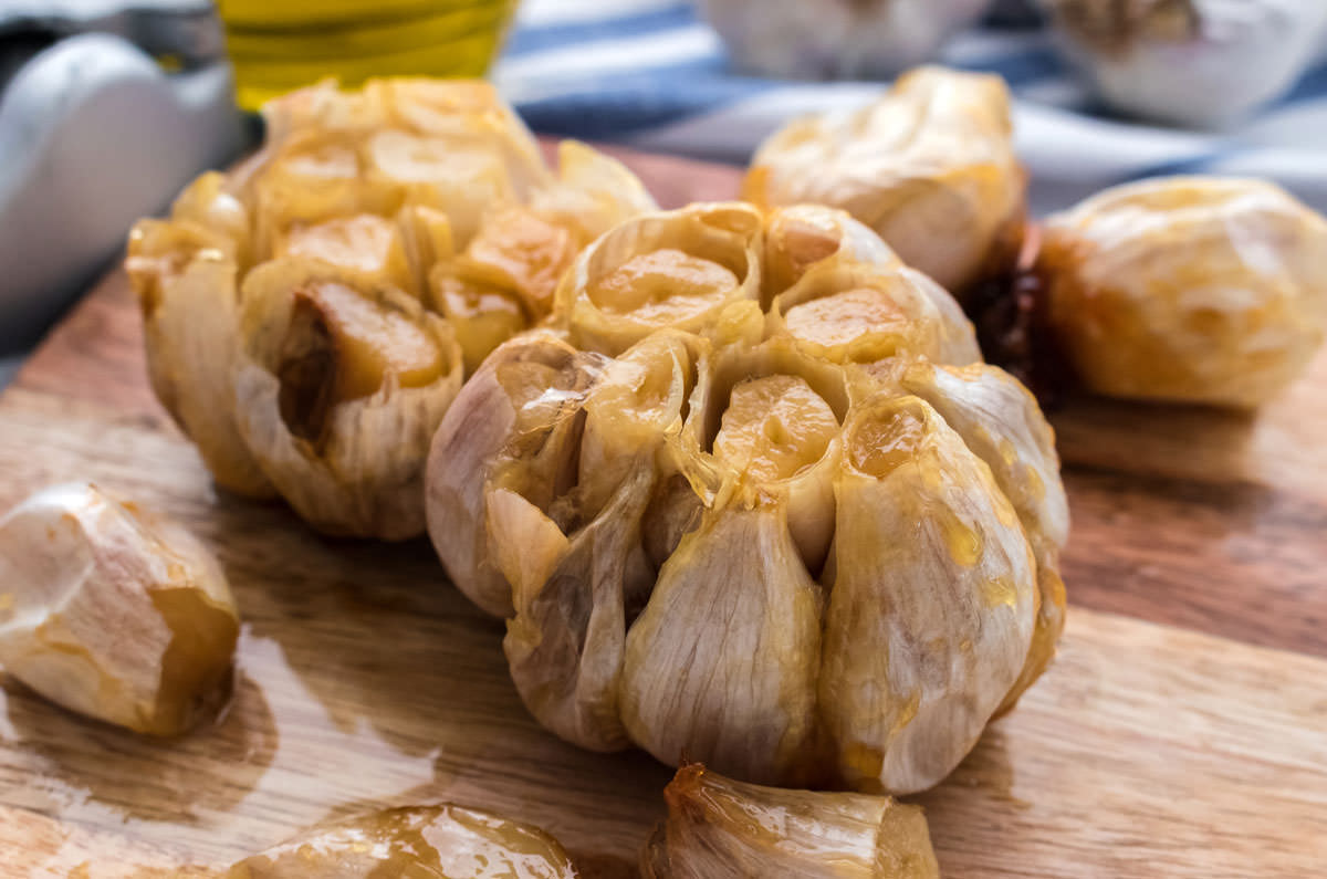 Heads of roasted garlic sitting on a cutting board in front of a blue and white towel, olive oil and a garlic press.
