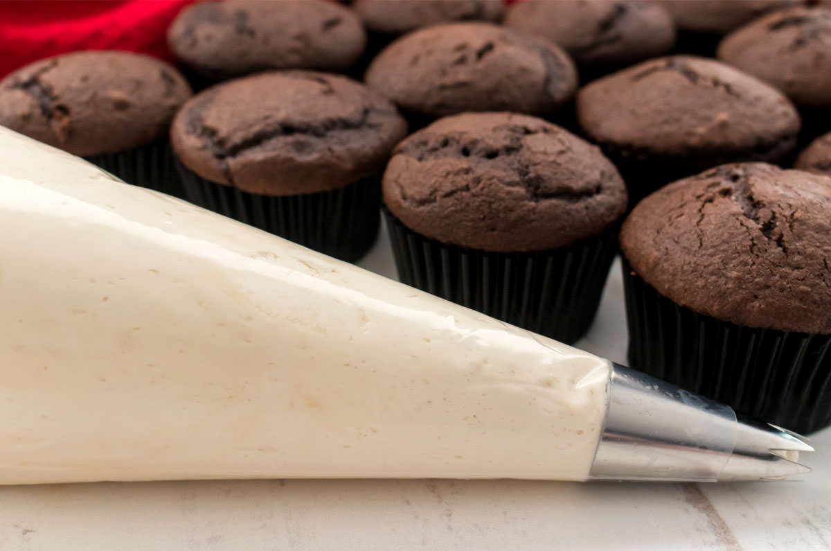 Closeup on a decorating bag filled with Kahlua Buttercream Frosting sitting on a white table with a batch of unfrosted cupcakes.