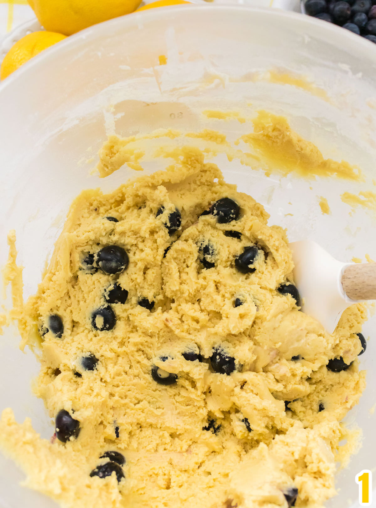 Closeup on a glass mixing bowl filled with Lemon Blueberry cookie dough and a white wooden spatula.