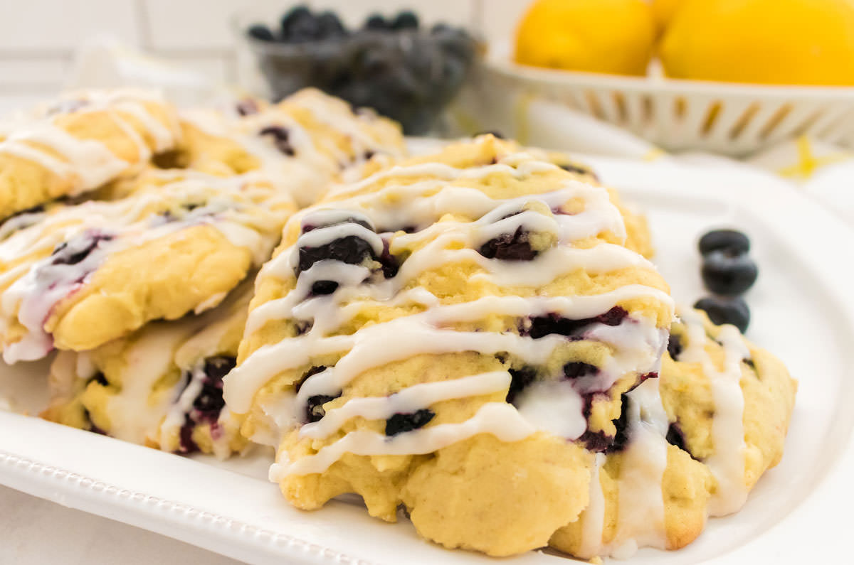 Closeup on a white platter filled with Lemon Blueberry Cookies sitting on a white table in front of a bowl of fresh blueberries and a bowl of fresh lemons.
