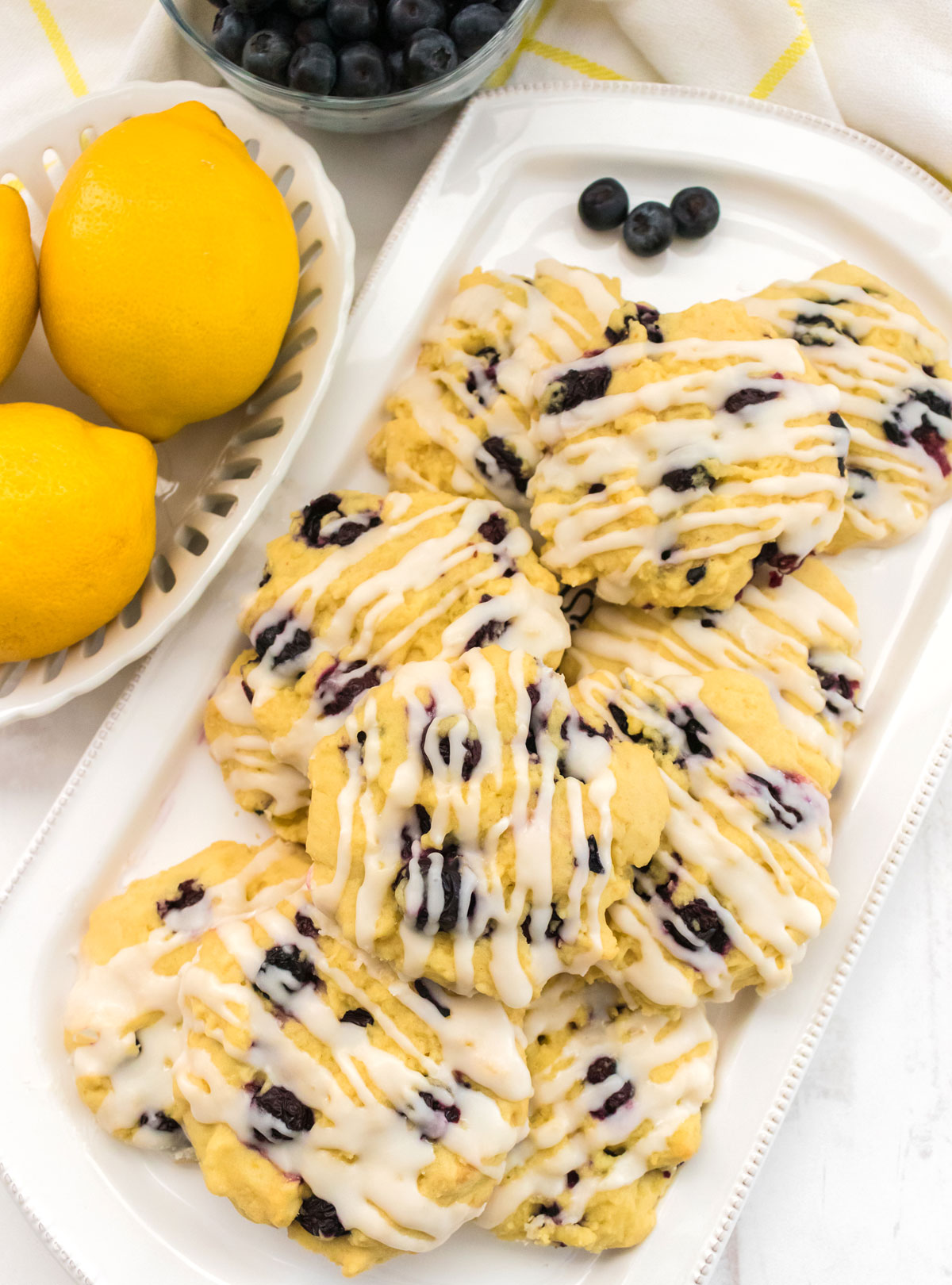 Overhead shot of a white serving platter filled with Lemon Blueberry Cookies sitting next to a bowl of lemons and a bowl of fresh blueberries.