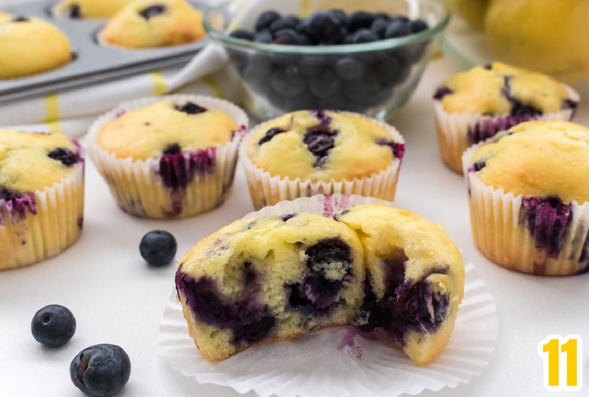 Closeup on six unfrosted Lemon Blueberry Cupcakes sitting on a white table with a bowl of lemons and blueberries behind them.