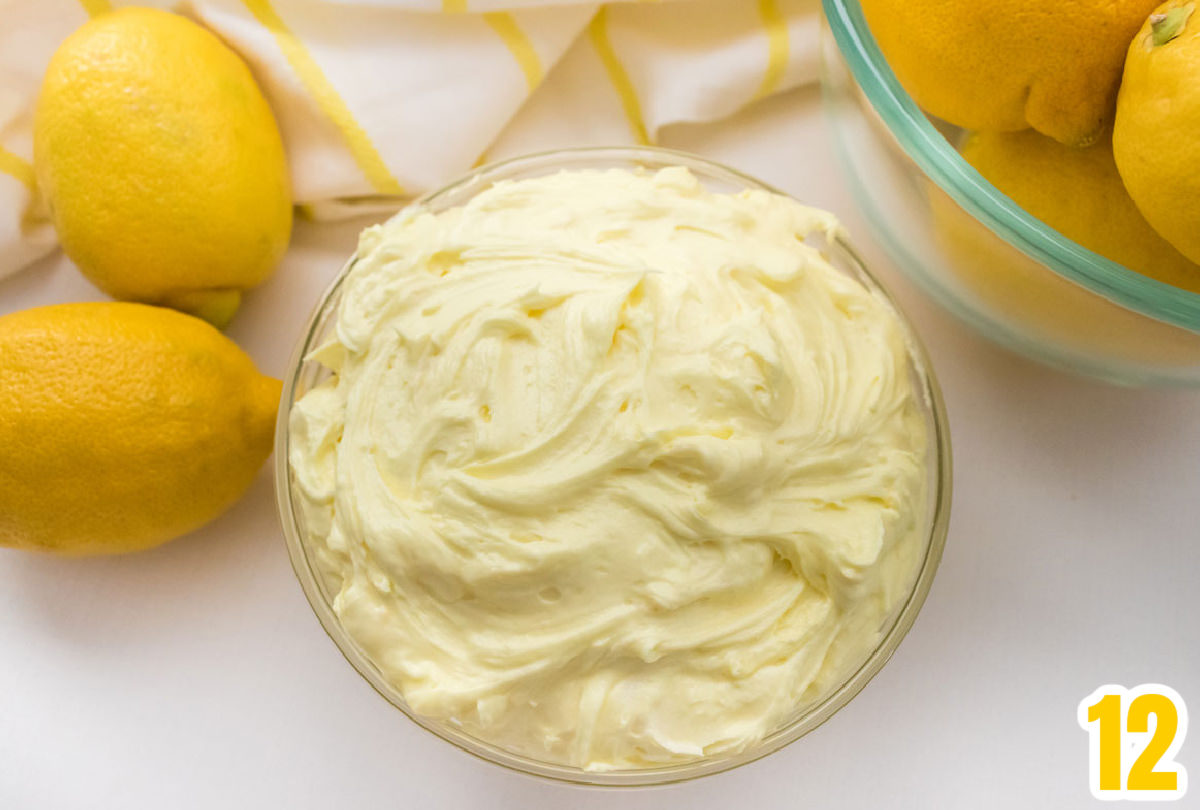 Closeup on a glass bowl filled with homemade Lemon Buttercream Frosting surrounded by lemons and a white and yellow towel.