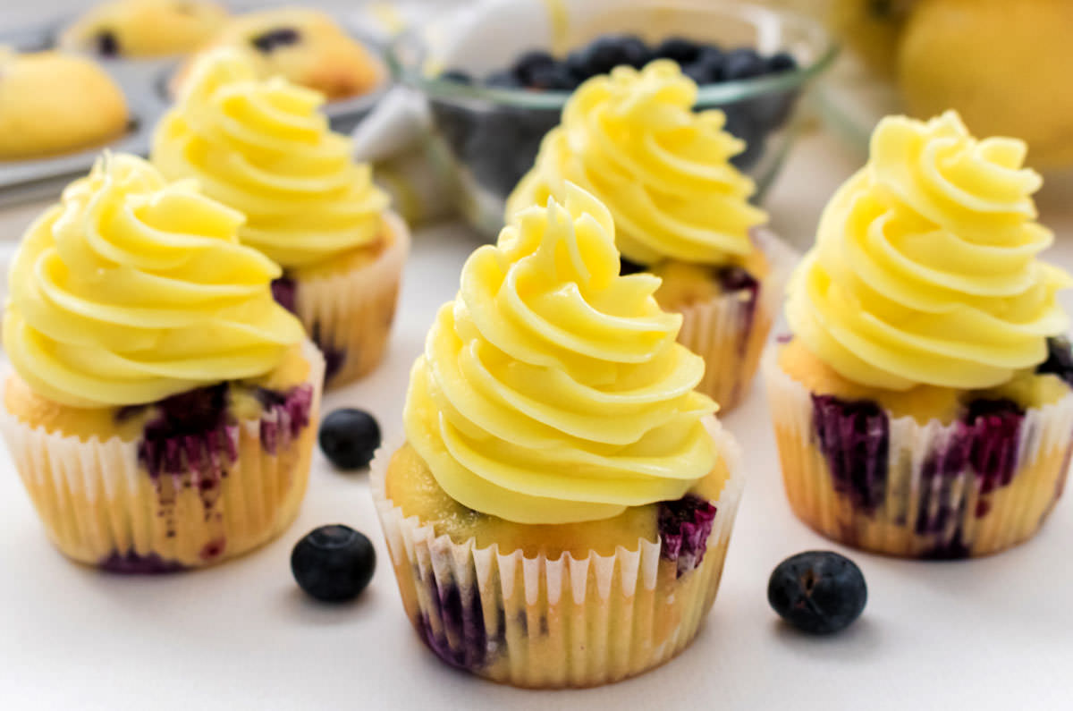 Closeup on five Lemon Blueberry Cupcakes with Lemon Buttercream Frosting sitting on a white surface surrounded by blueberries, lemons and a tin of cupcakes.