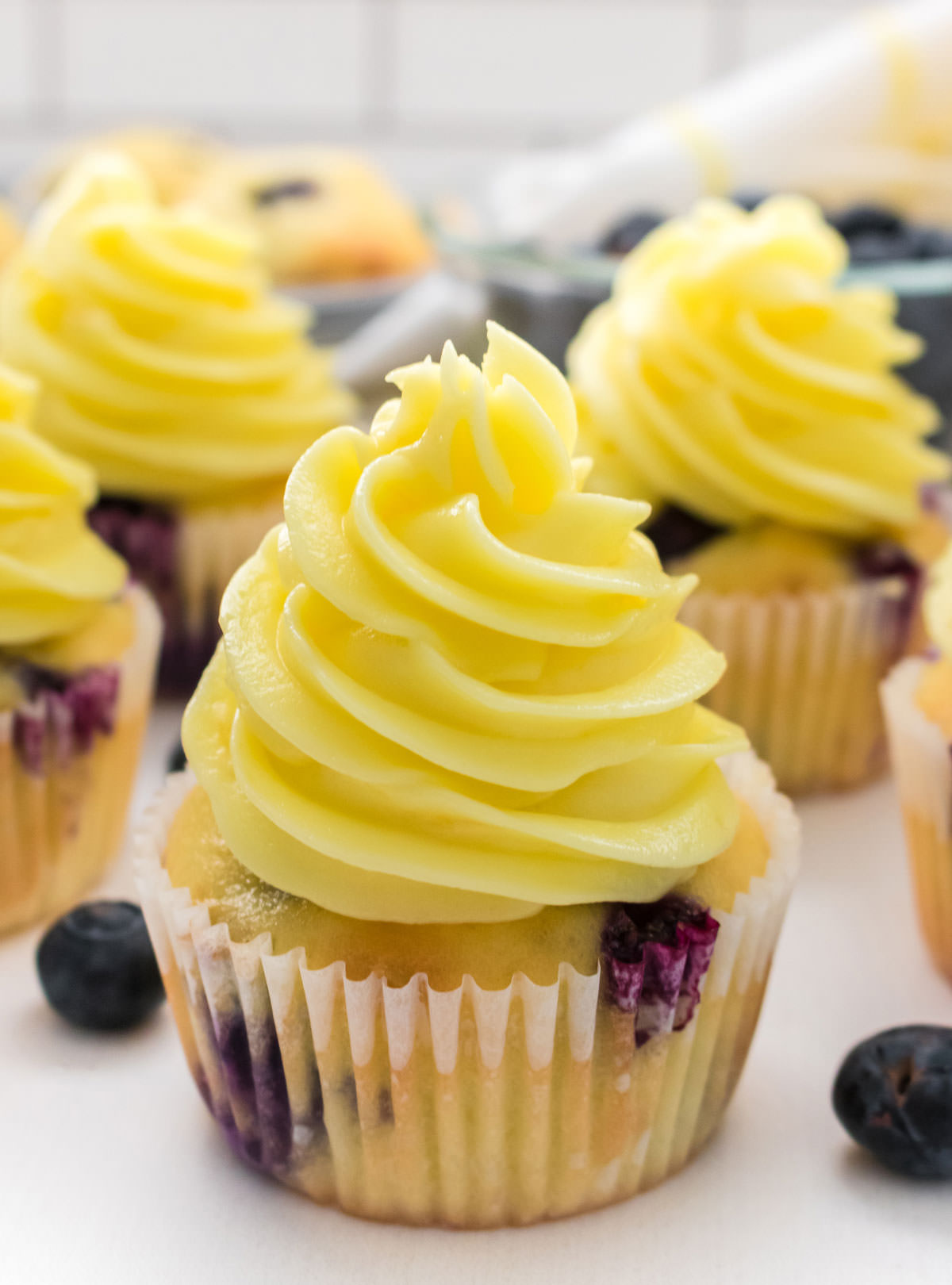 Closeup of a Lemon Blueberry Cupcake with Lemon Frosting sitting on a white table surrounded by other cupcakes and fresh blueberries.