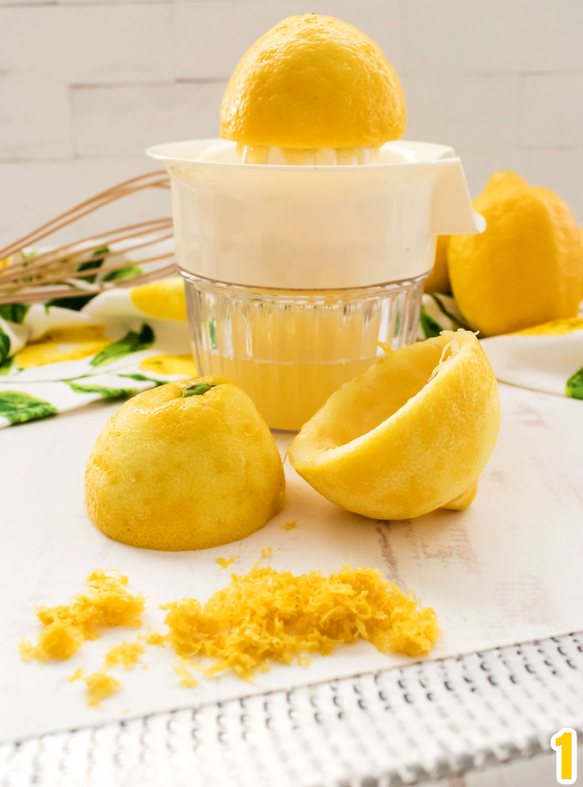 Close up of a Lemon Juicer filled with lemon juice, a microplane that zested the lemons, a whisk and some fresh lemons.