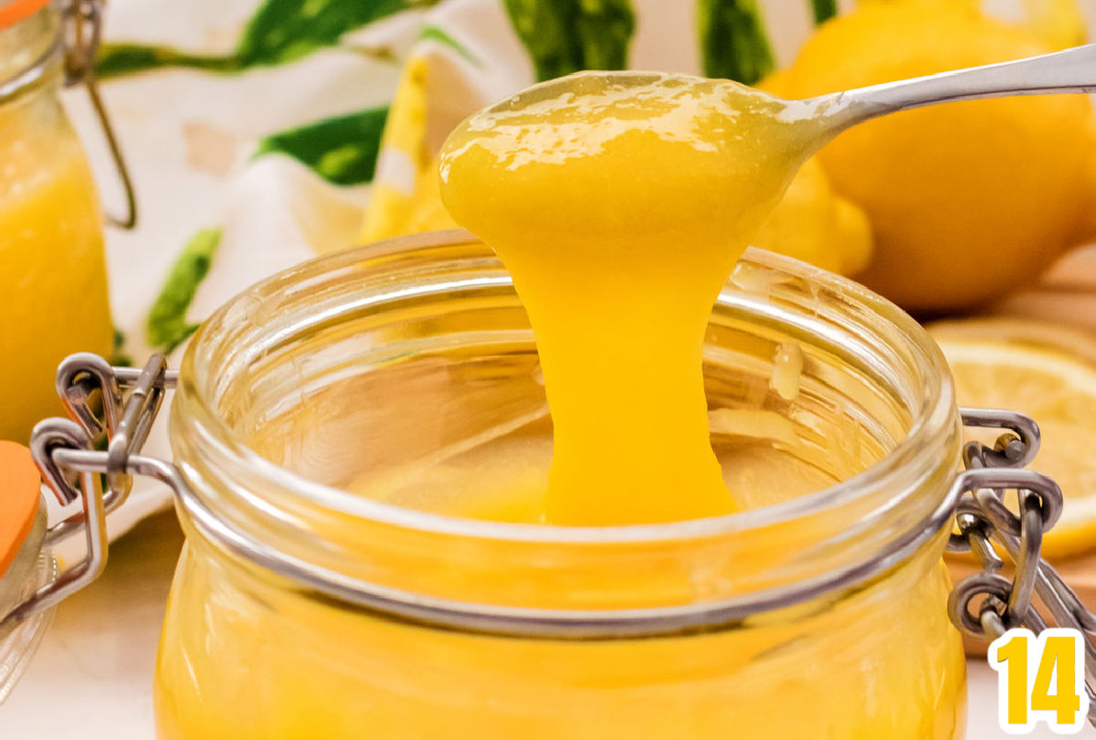 Closeup on a spoon filled with Lemon Curd hovering over a glass jar filled with our Easy Lemon Curd with lemons and a second jar of lemon curd in the background.