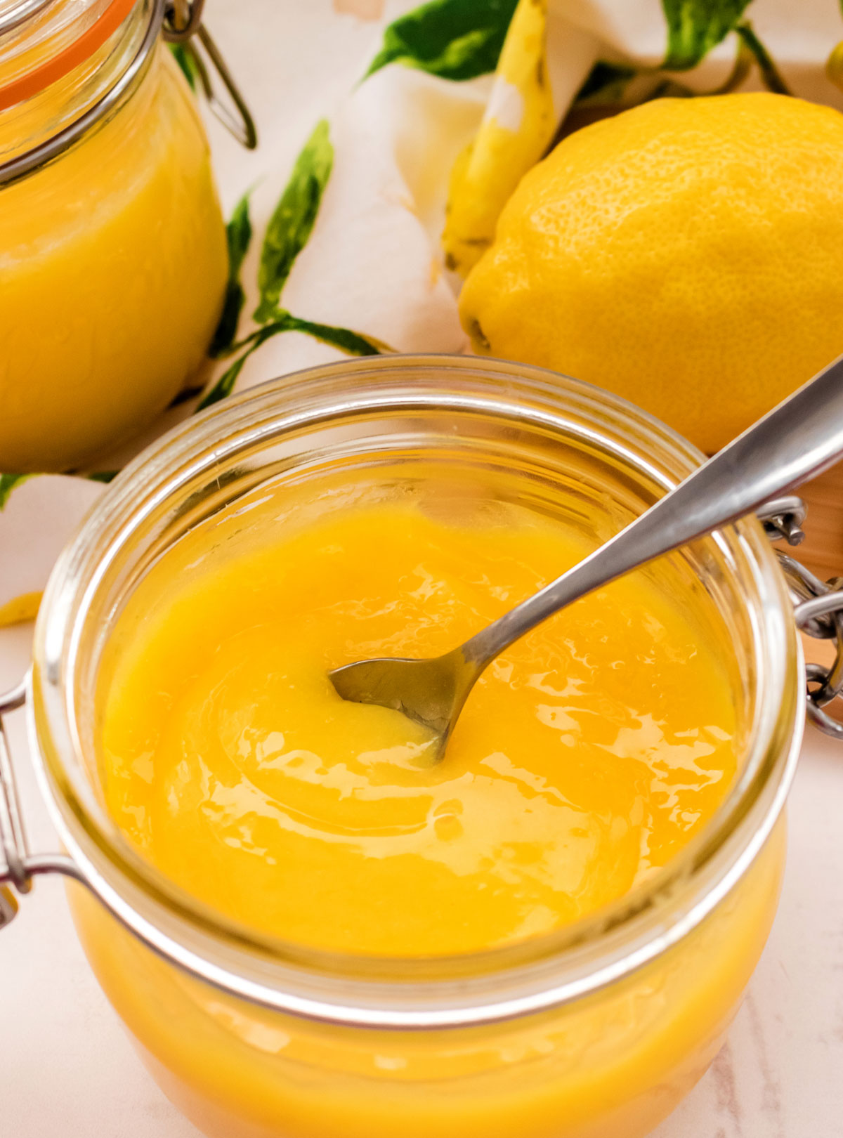 A glass jar filled with Homemade Lemon Curd and a sliver spoon surrounded by fresh lemons and a kitchen towel.