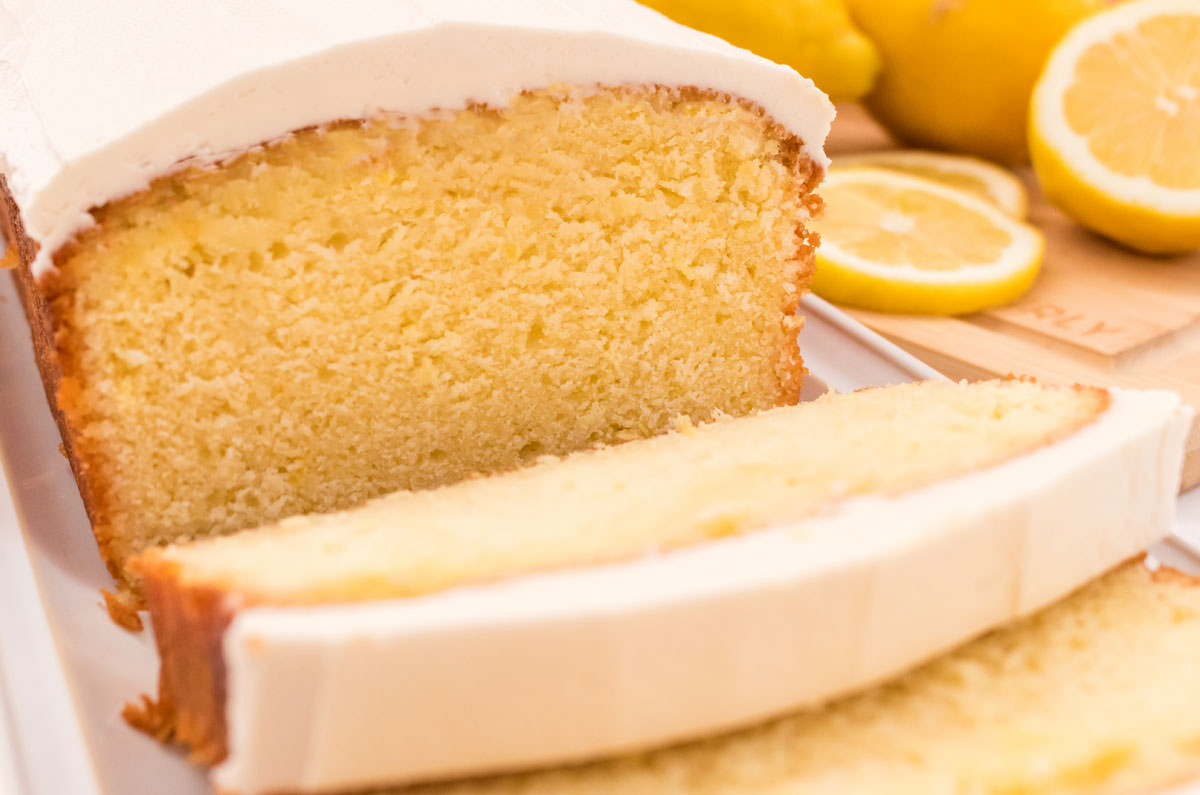 Closeup on a Lemon Pound Cake frosted with Vanilla Icing sitting on a white plate next to a cutting board covered with lemon slices.