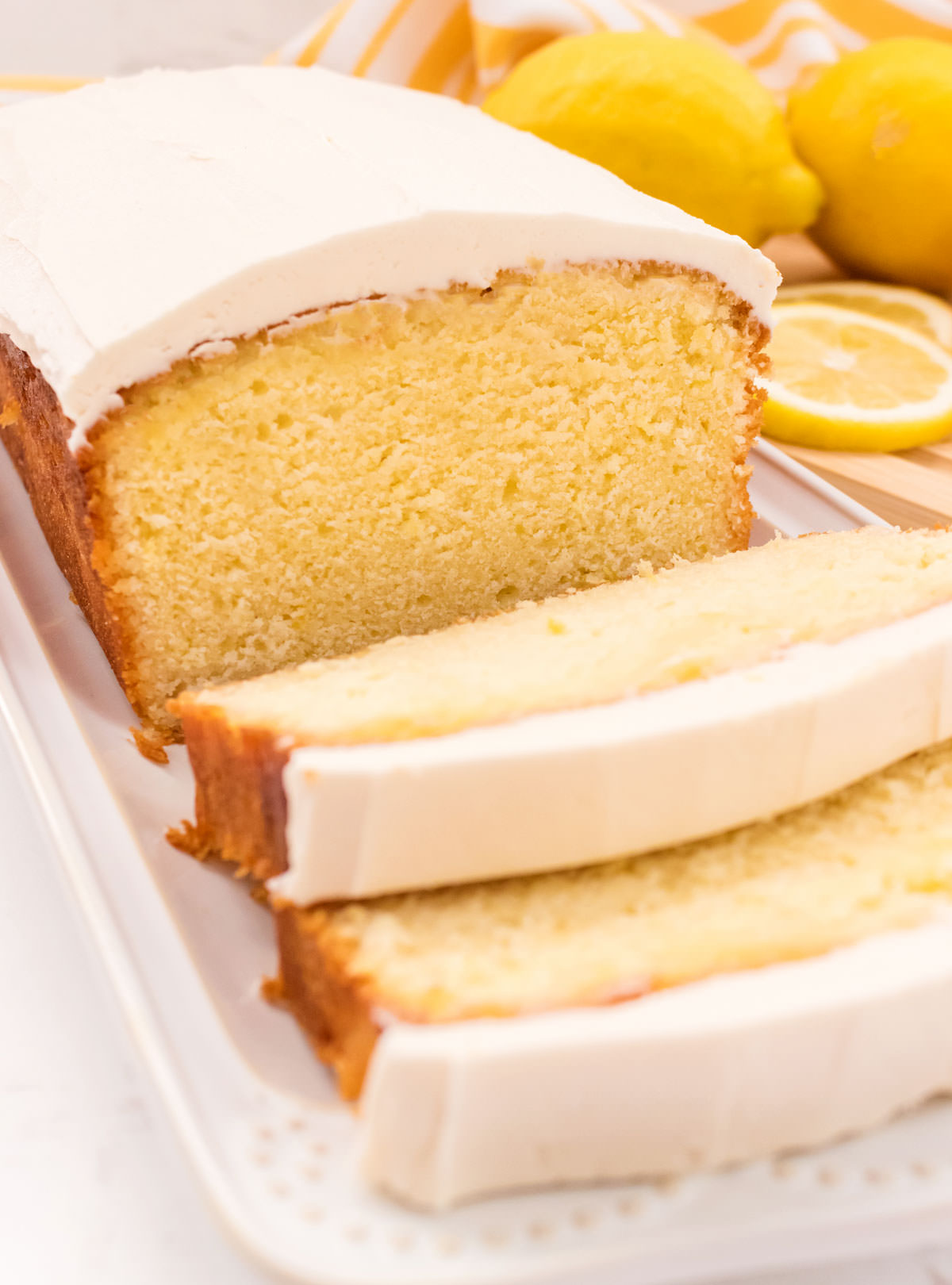 Closeup on a partially sliced Lemon Pound Cake sitting on a white serving platter on a white table with lemons in the background.