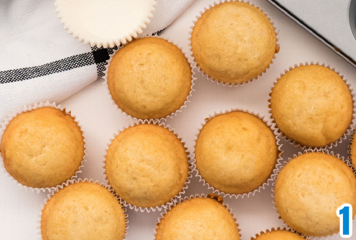 A dozen vanilla cupcakes sitting on a white table in front of a cupcake tin and a black and white towel.