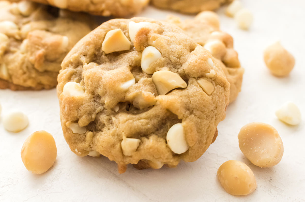 Closeup on a White Chocolate Macadamia Nut Cookie sitting on a white table surrounded by White Chocolate Chips.