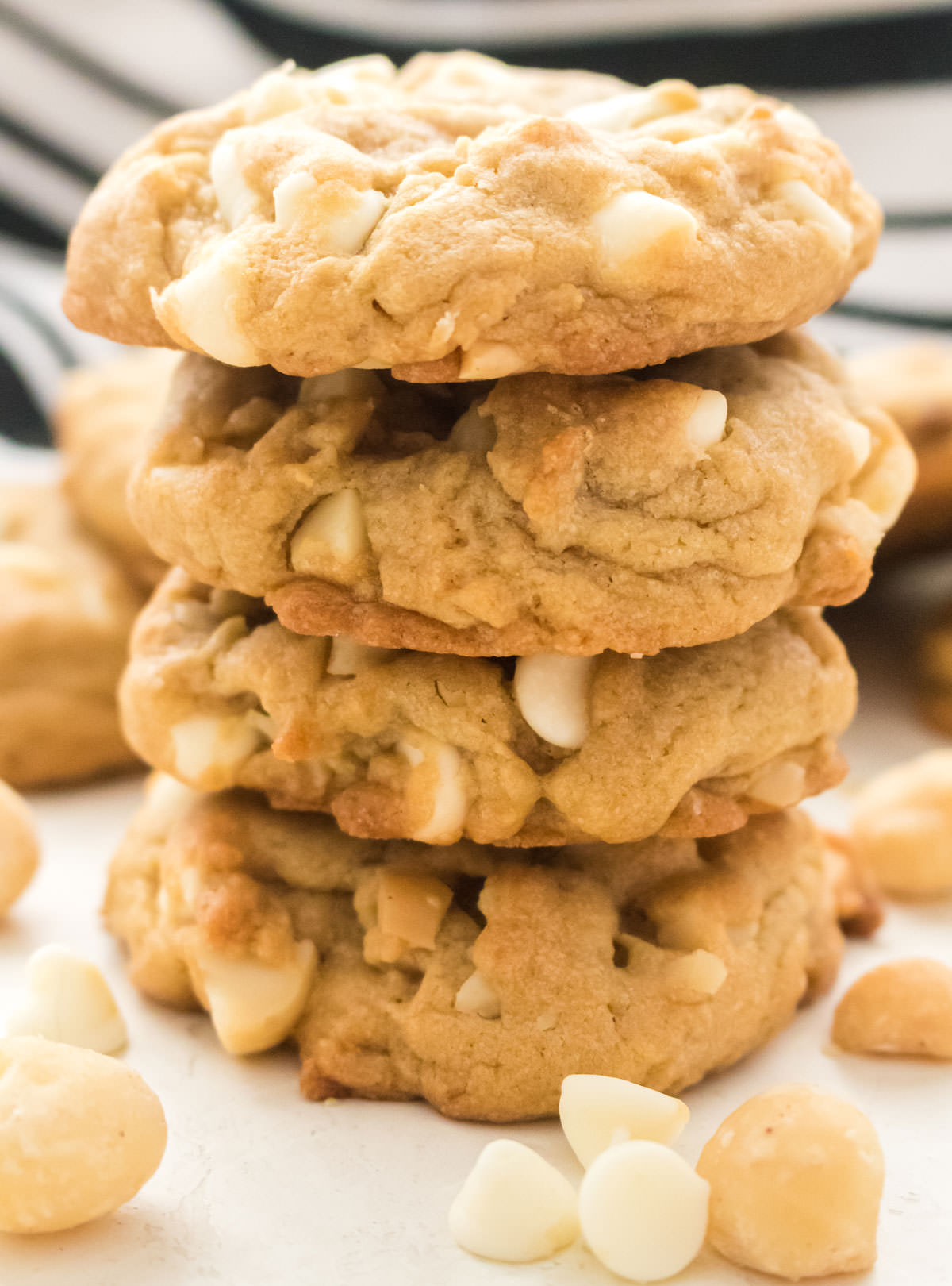 Closeup on a stack of White Chocolate Macadamia Nut Cookies sitting on a white table surrounded by White Chocolate Chips.