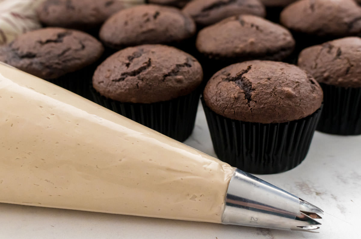 Closeup on a decorating bag filled with Maple Frosting sitting in front of a batch of unfrosted chocolate cupcakes.