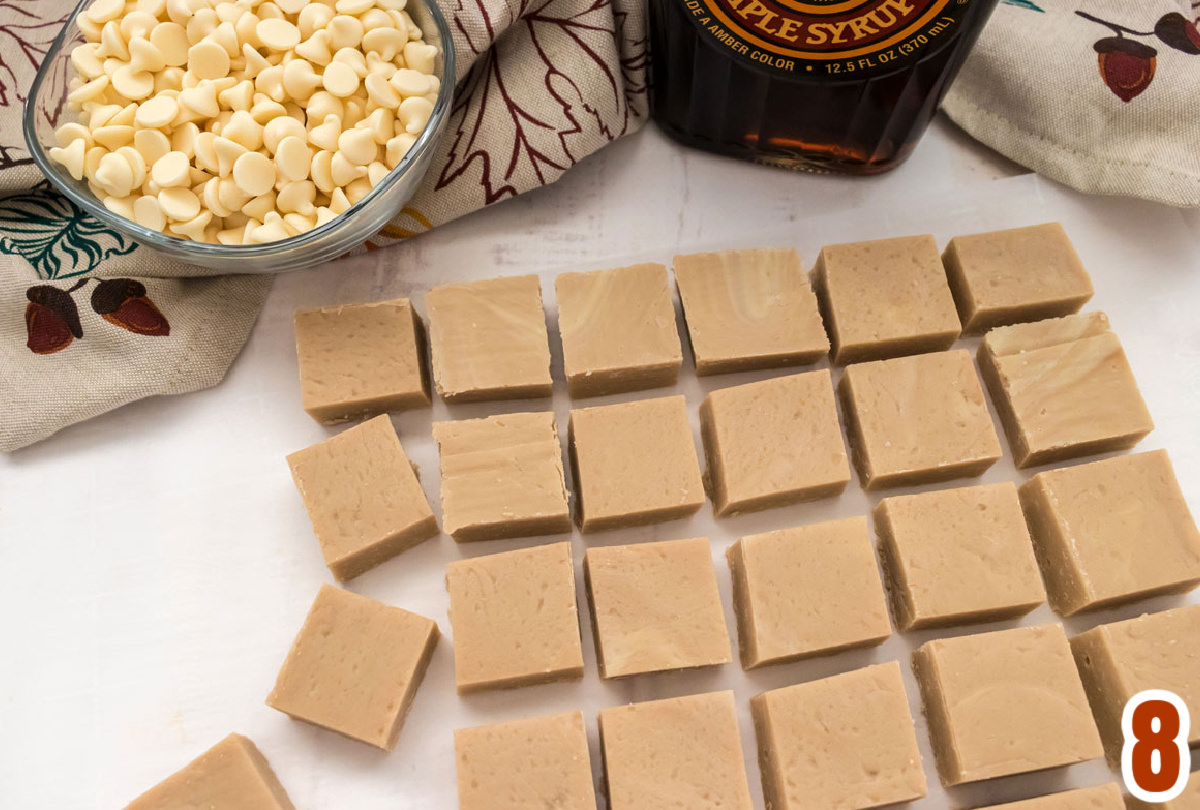 Overhead shot of a batch of Maple Fudge lined up in rows on a white table in front of a bottle of Maple Syrup and a fall table linen.