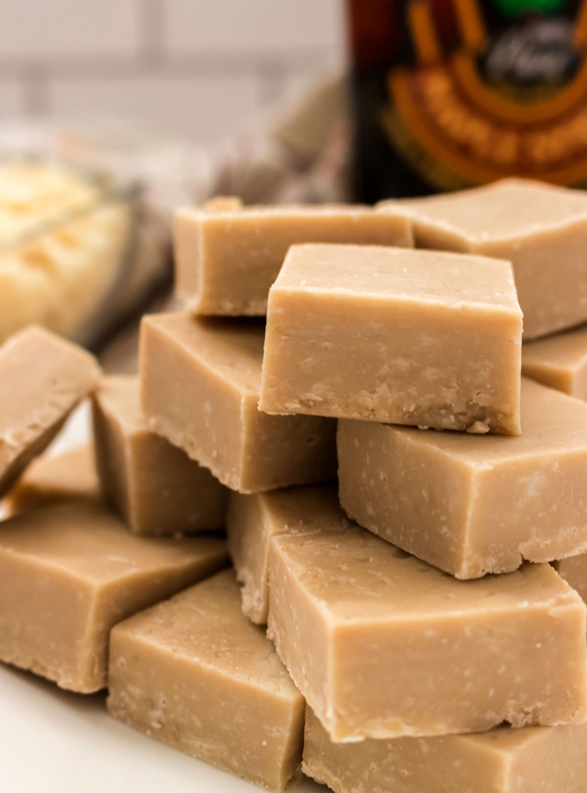 Closeup on a stack of Maple Fudge sitting on a white surface in front of a bottle of maple syrup and a glass bowl filled with White Chocolate Chips.