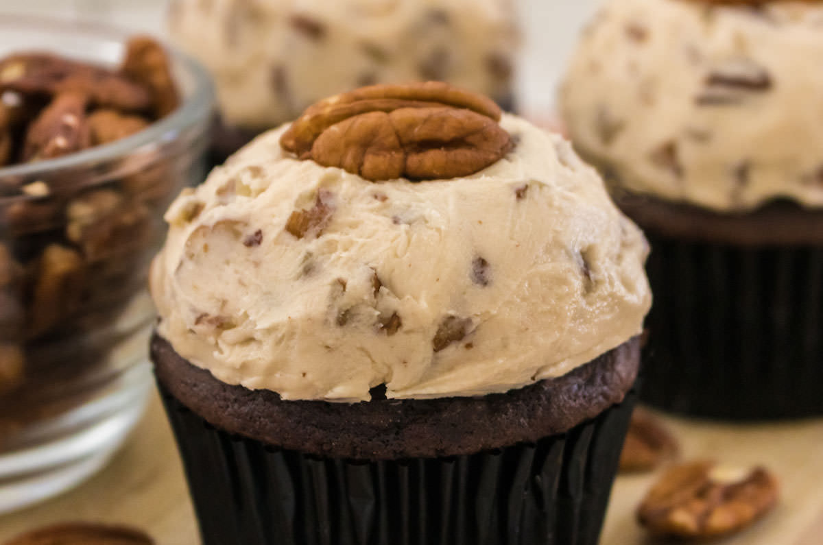 Closeup on a cupcake topped with Maple Pecan Buttercream Frosting sitting next to a glass bowl filled with pecans.
