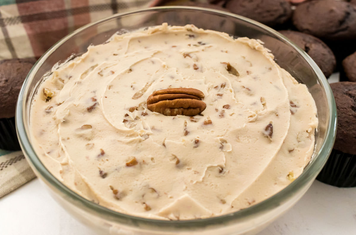 Closeup on a glass bowl filled with Maple Pecan Frosting sitting next to a batch of unfrosted chocolate cupcakes.