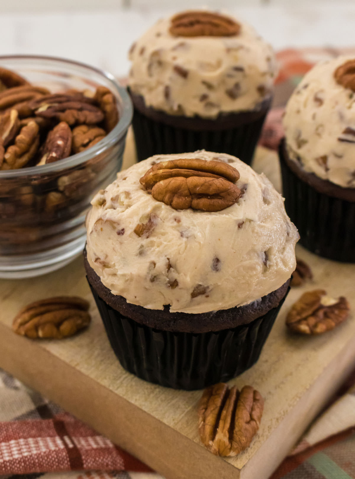 Closeup on three chocolate cupcakes topped with Maple Pecan Buttercream Frosting sitting on a wooden cutting board next to a glass bowl filled with pecans.