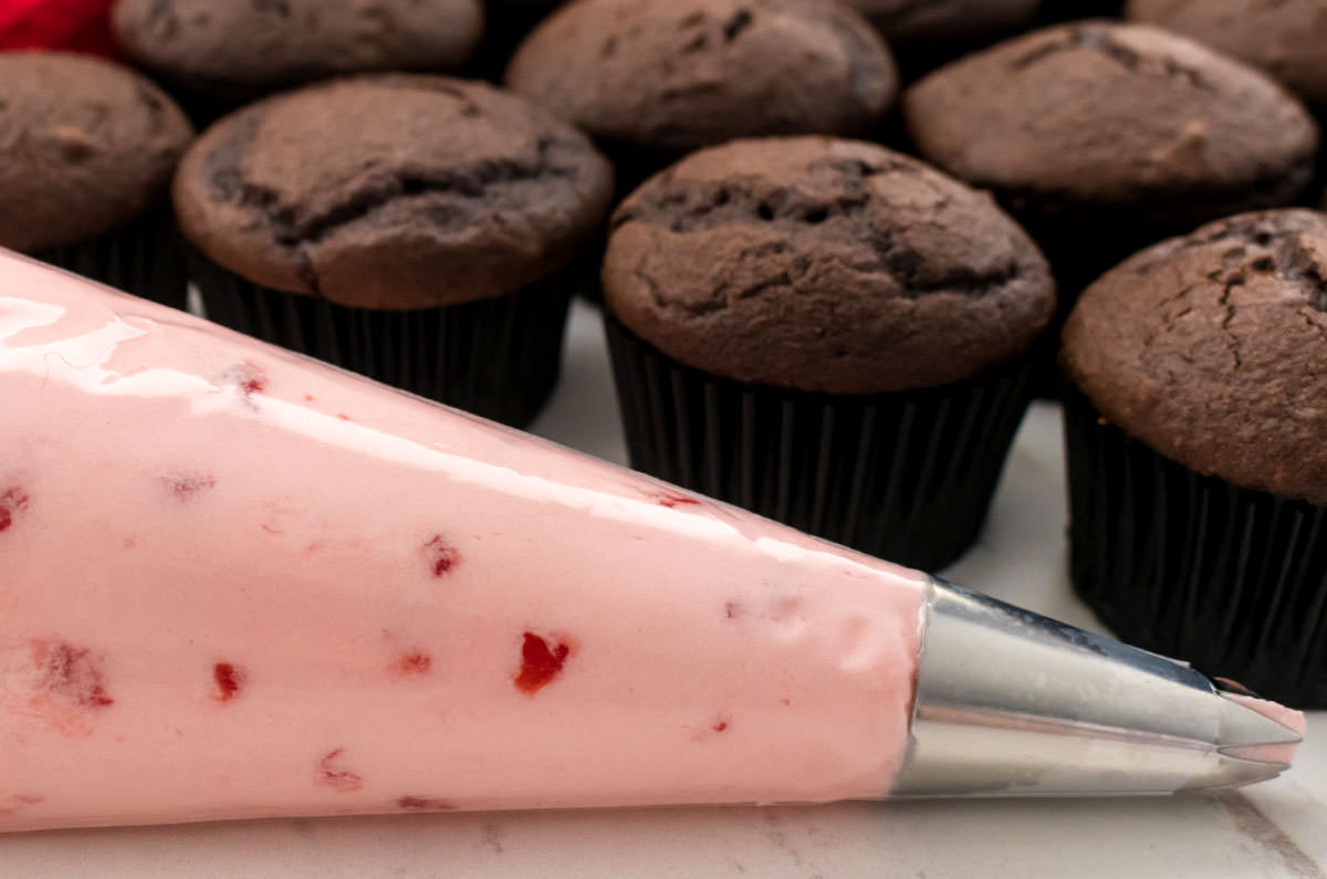 Closeup on a decorating bag filled with Cherry Frosting sitting in front of a batch of unfrosted chocolate cupcakes.