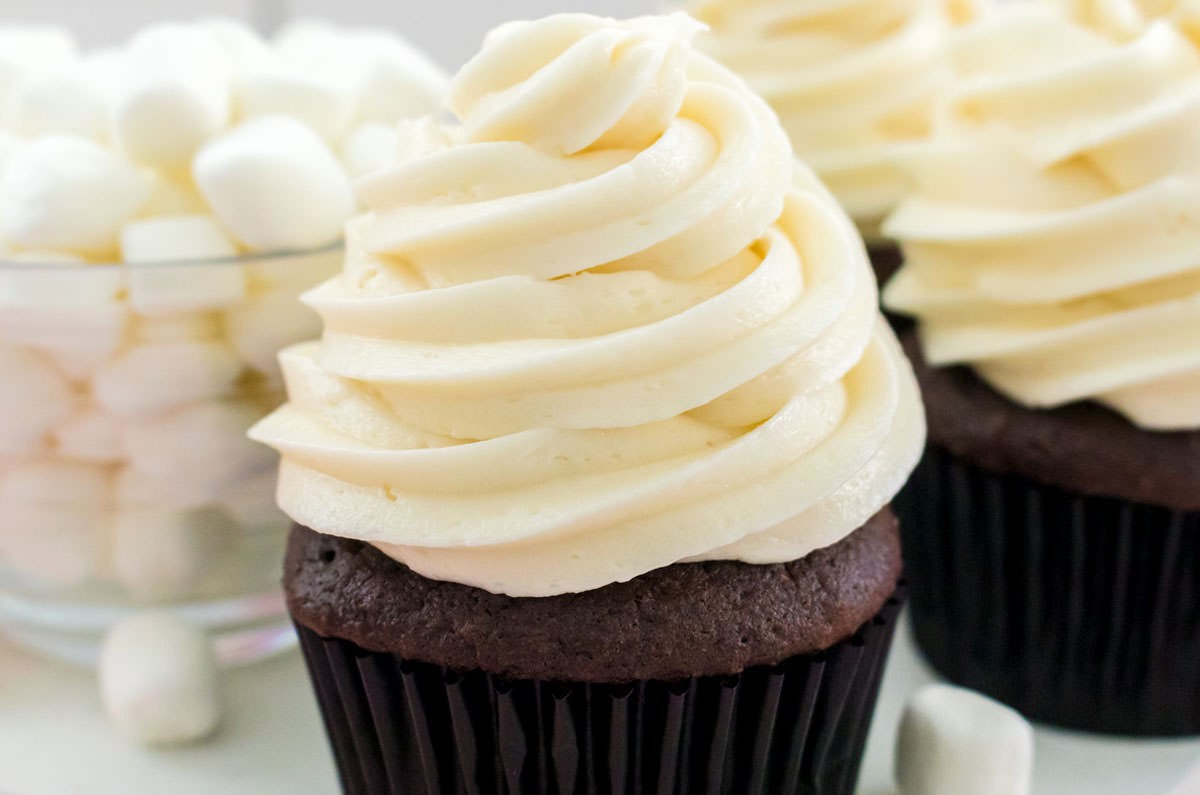 Closeup on three chocolate cupcakes topped with The Best Marshmallow Frosting sitting next to a glass bowl filled with Mini Marshmallows.