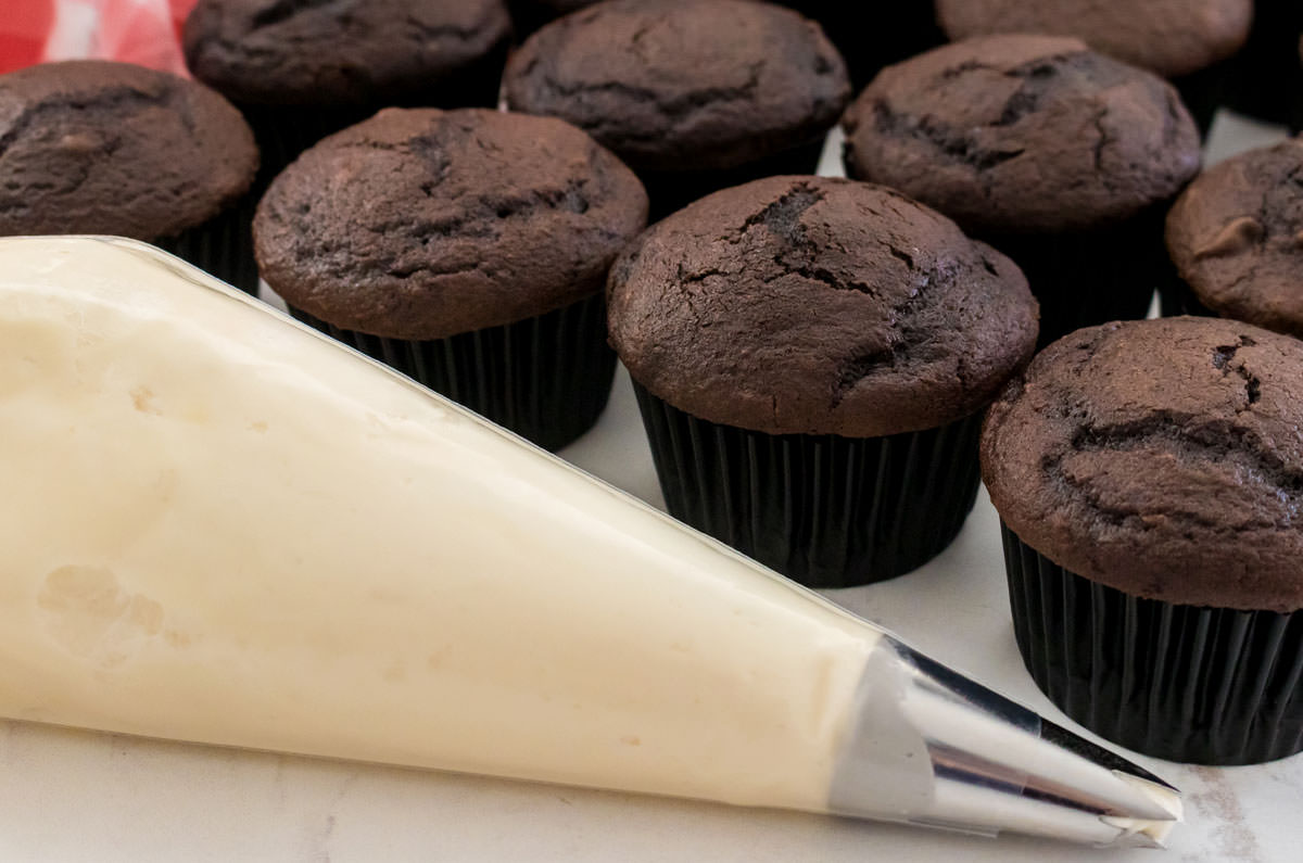 Decorating bag filled with Marshmellow Frosting sitting on a white table next to a batch of unfrosted chocolate cupcakes.