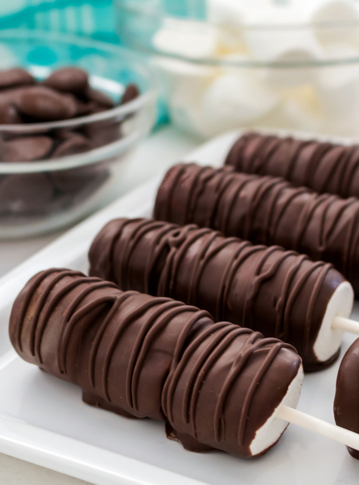 Close up on a white platter holding four Marshmallow Pops sitting in front of a glass bowl filled with chocolate and another glass bowl filled with marshmallows.