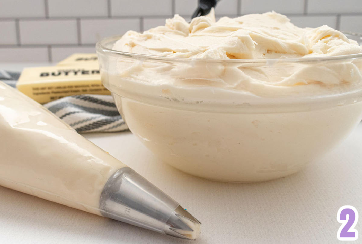 Closeup on a glass bowl filled with buttercream frosting sitting next to sticks of butter and a decorating bag filled with frosting.