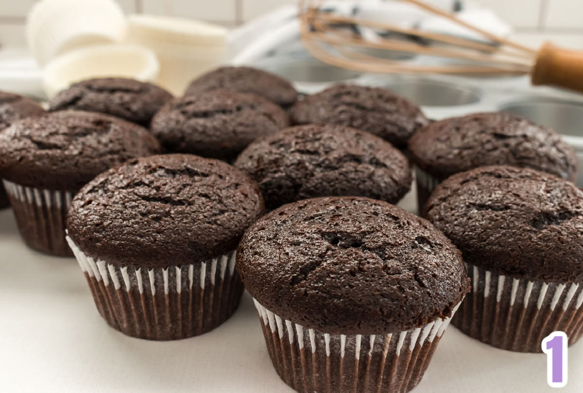 Ten chocolate cupcakes sitting on a white table in front of a cupcake tin and white cupcake liners.