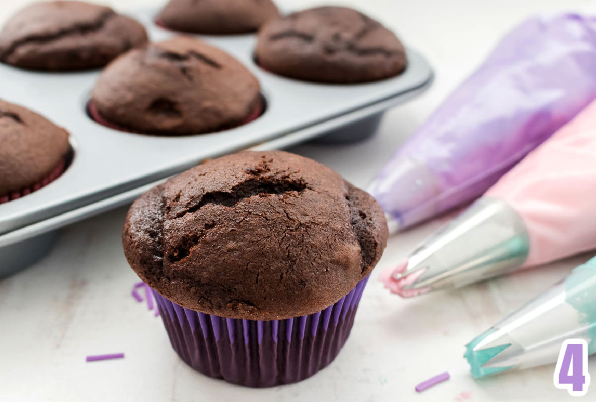 Chocolate cupcake in a purple cupcake liner sitting in front of a tin of cupcakes and buttercream frosting in decorating bags.