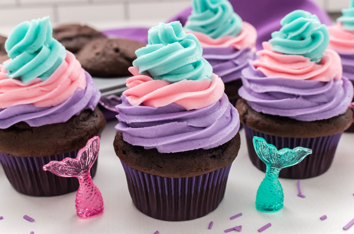 Closeup on four Mermaid Cupcakes sitting on a white table in front of a tin filled with cupcakes and a purple table linen.