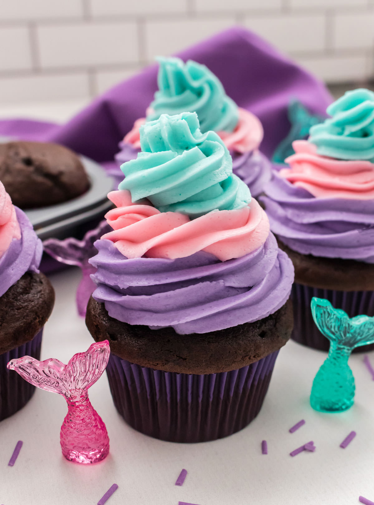 Closeup on three Mermaid Cupcakes sitting on a white table with colored Mermaid Tails surrounding it.