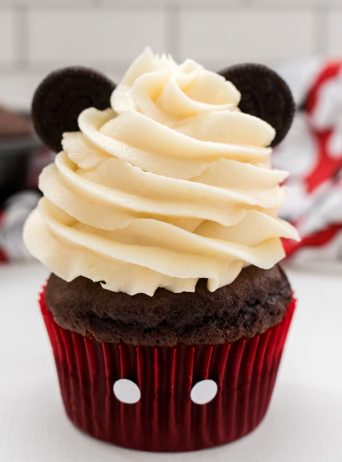 Closeup on a Mickey Mouse Cupcake sitting on a white table in front of a white background, Mickey Mouse Kitchen towel and a pan of cupcakes.