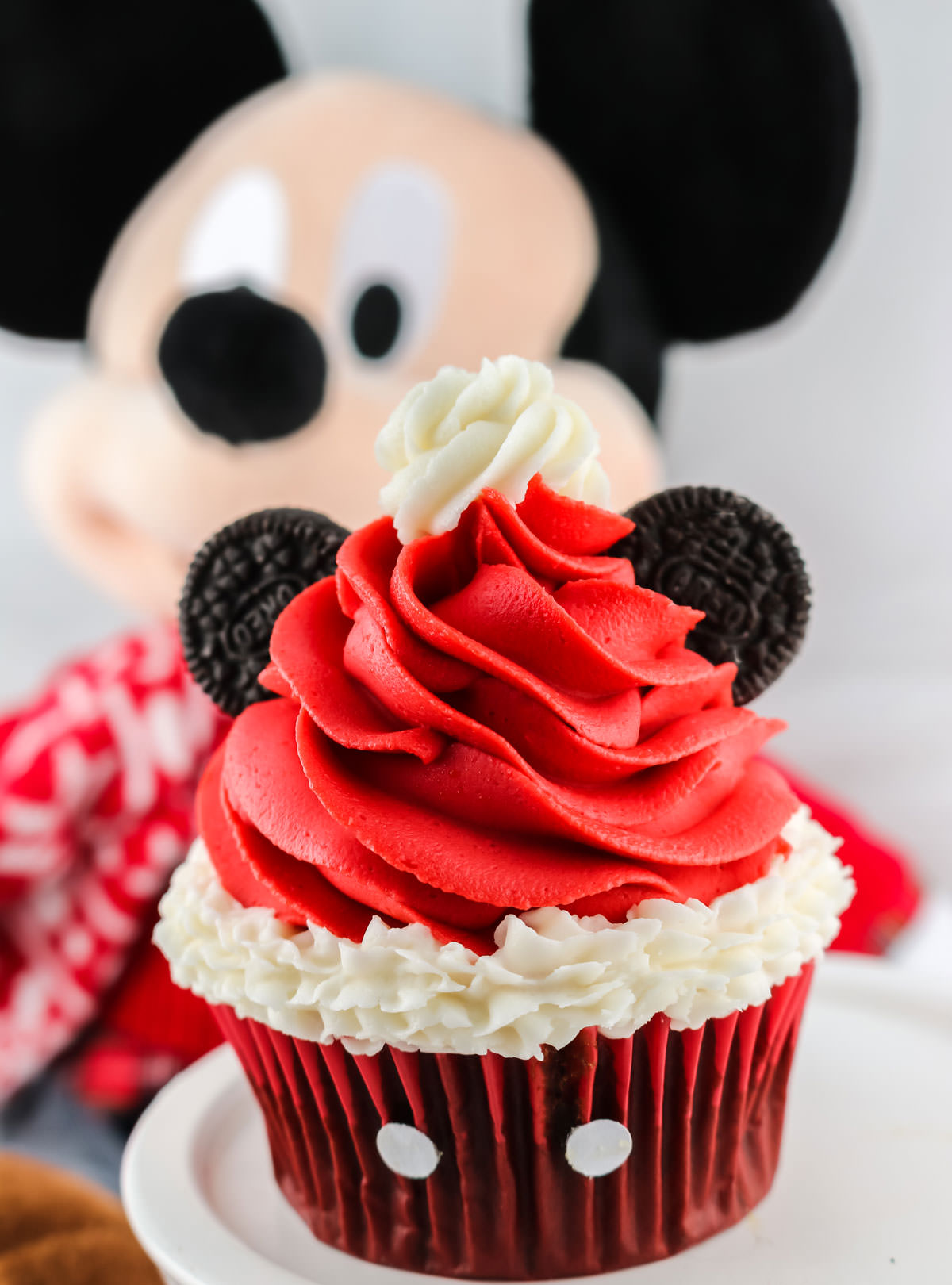 Closeup on a Mickey Mouse Santa Hat Cupcake sitting on a white cake plate in front of a Mickey Mouse stuffed animal.