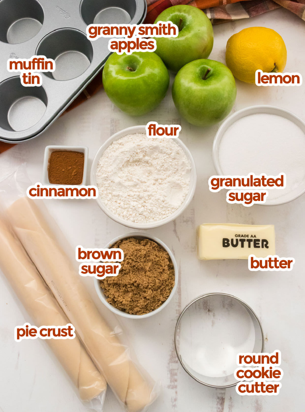 All the ingredients you will need to make Mini Apple Pies including Granny Smith Apples, lemon, flour. granulated sugar, cinnamon butter, brown sugar and pie crust.
