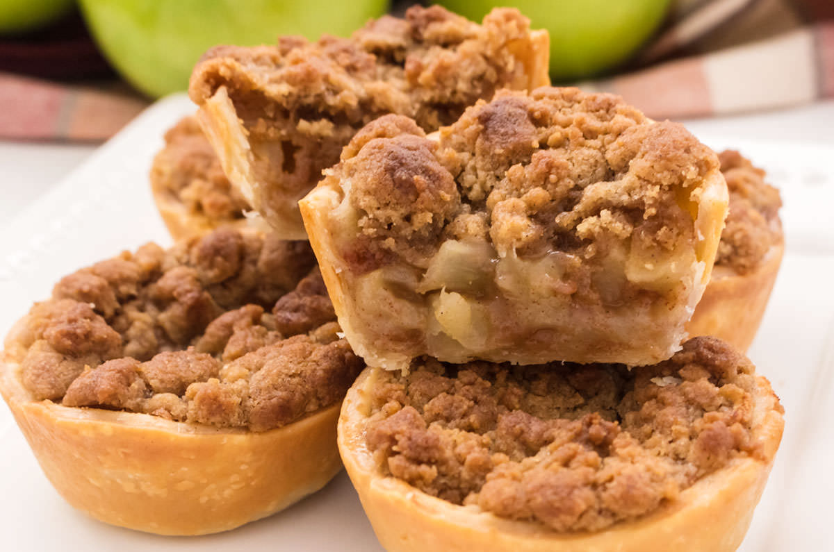 Closeup on five Mini Apple Pies sitting on a white plate in front of a stack of green apples.