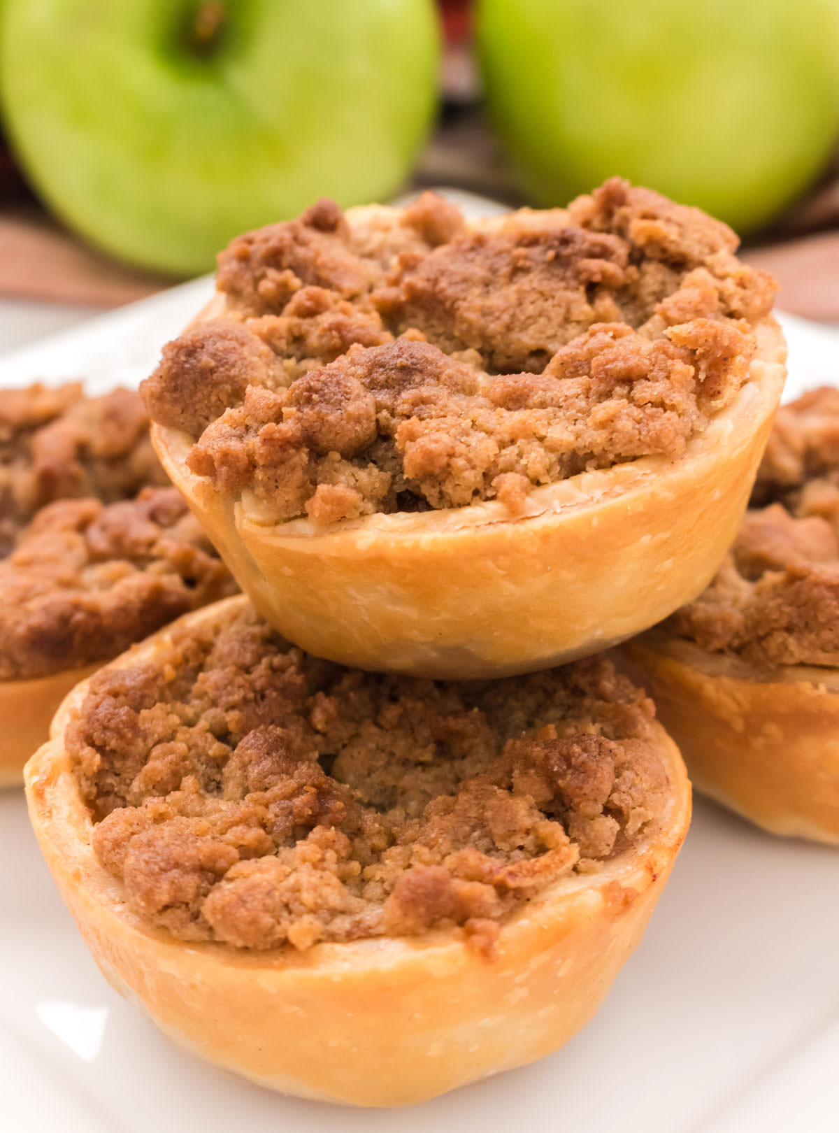 Closeup on five Mini Apple Pies sitting on a white plate in front a stack of green apples.