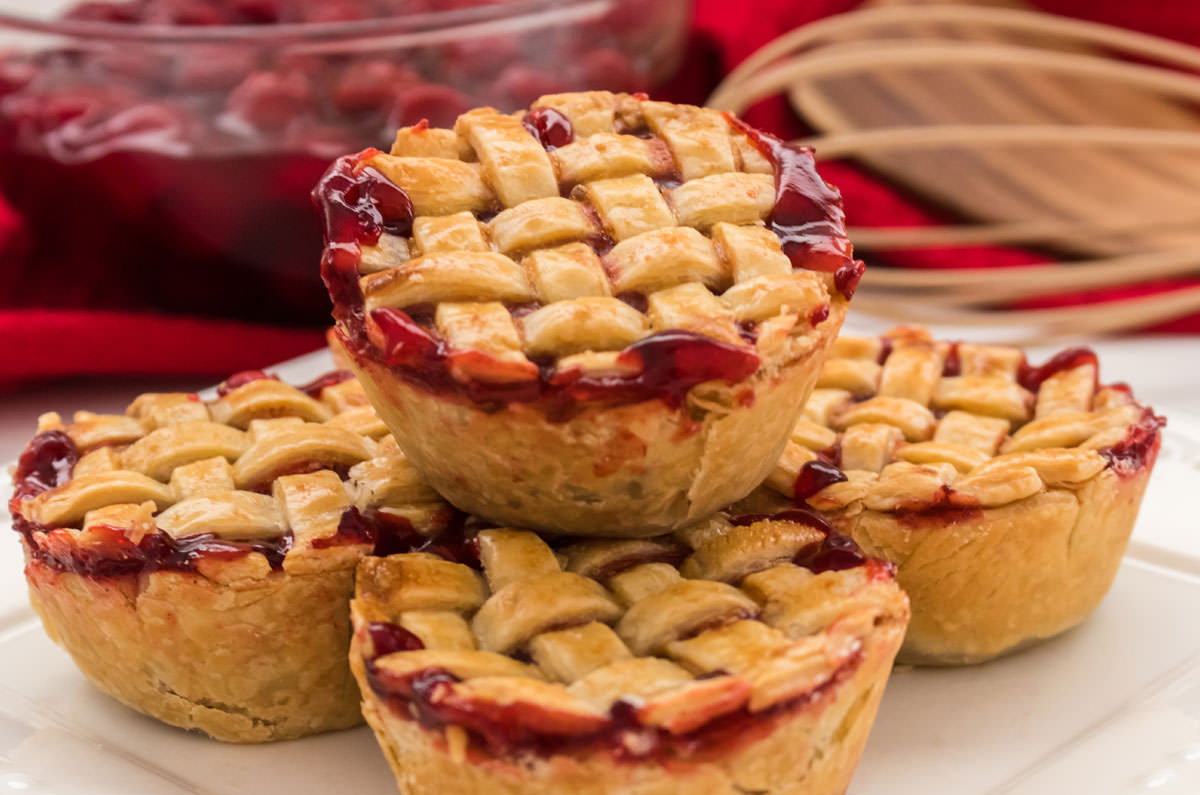 Closeup on a stack of five Mini Cherry Pies sitting on a white plate in front of a bowl filled with Cherry pie filling.