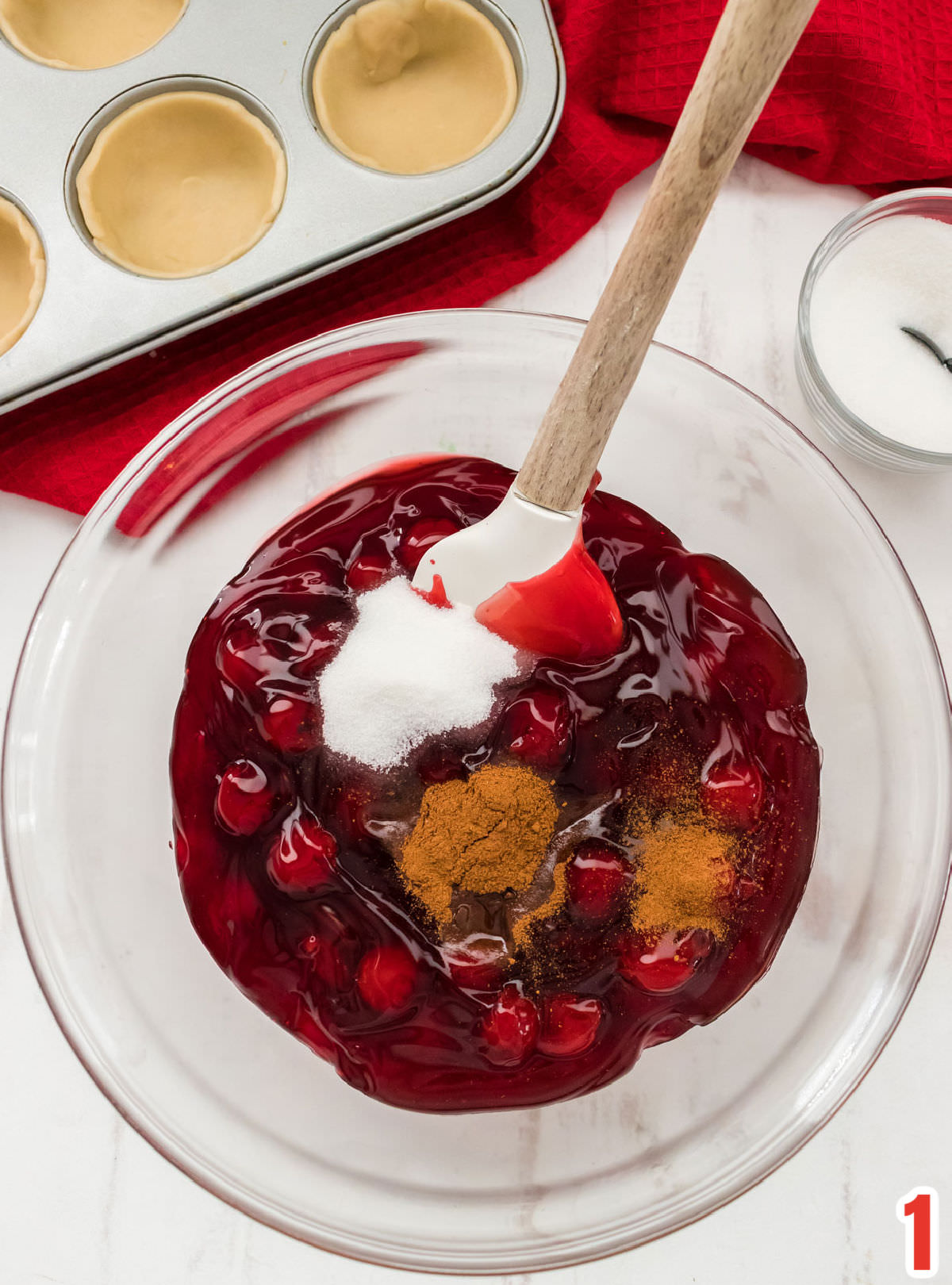 Closeup on a glass bowl filled with canned cherry pie filling, sugar, cinnamon and almond extract.