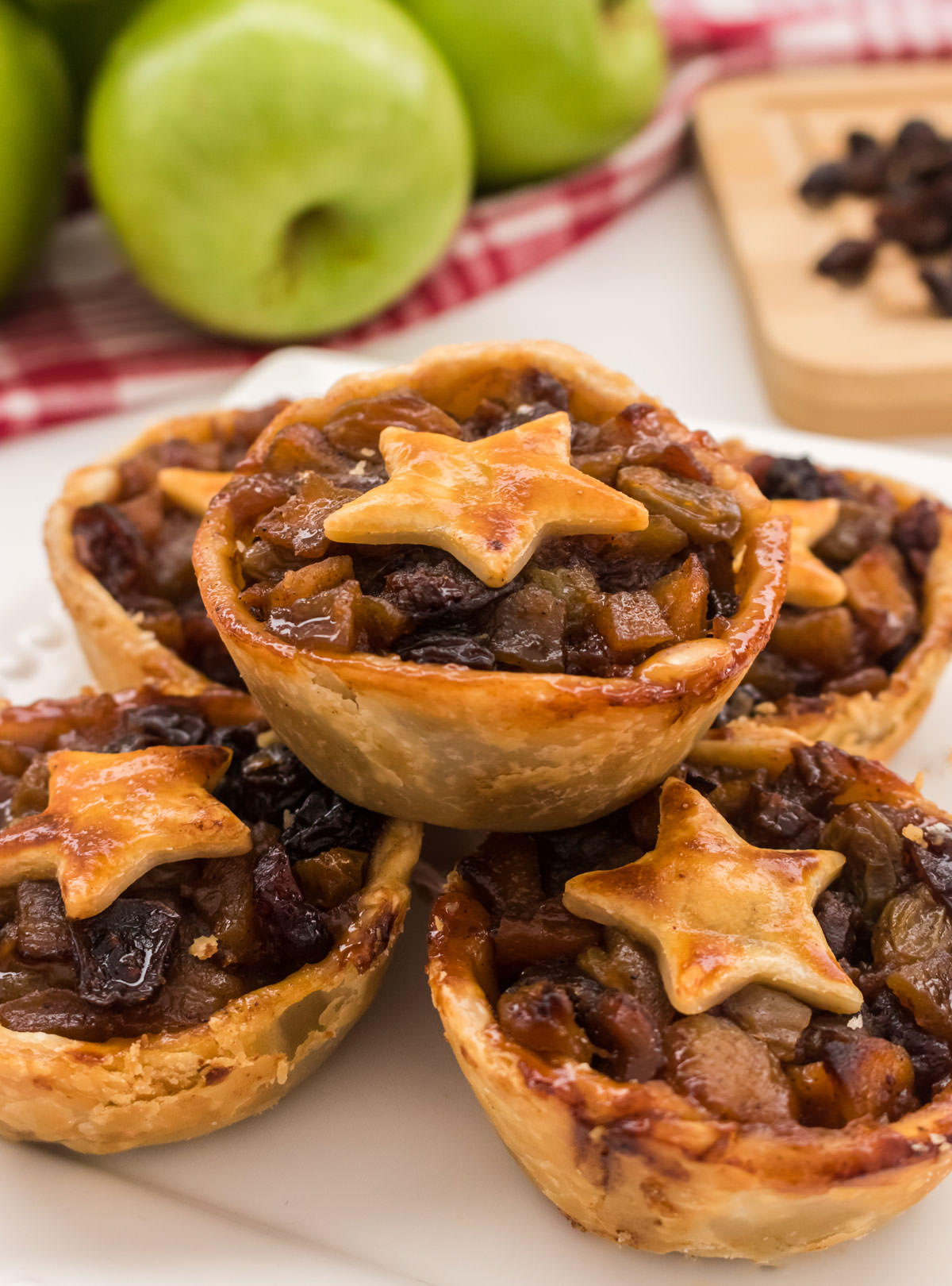 Closeup on five Mini Mince Pies sitting on a white plate in front of a stack of green apples.